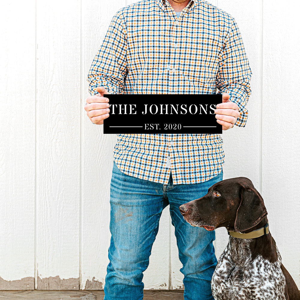 Man holding a personalized metal sign with 'The Johnsons Est. 2020' next to a dog against a white wooden background. The name and est date are customizable by Funlifenow.com