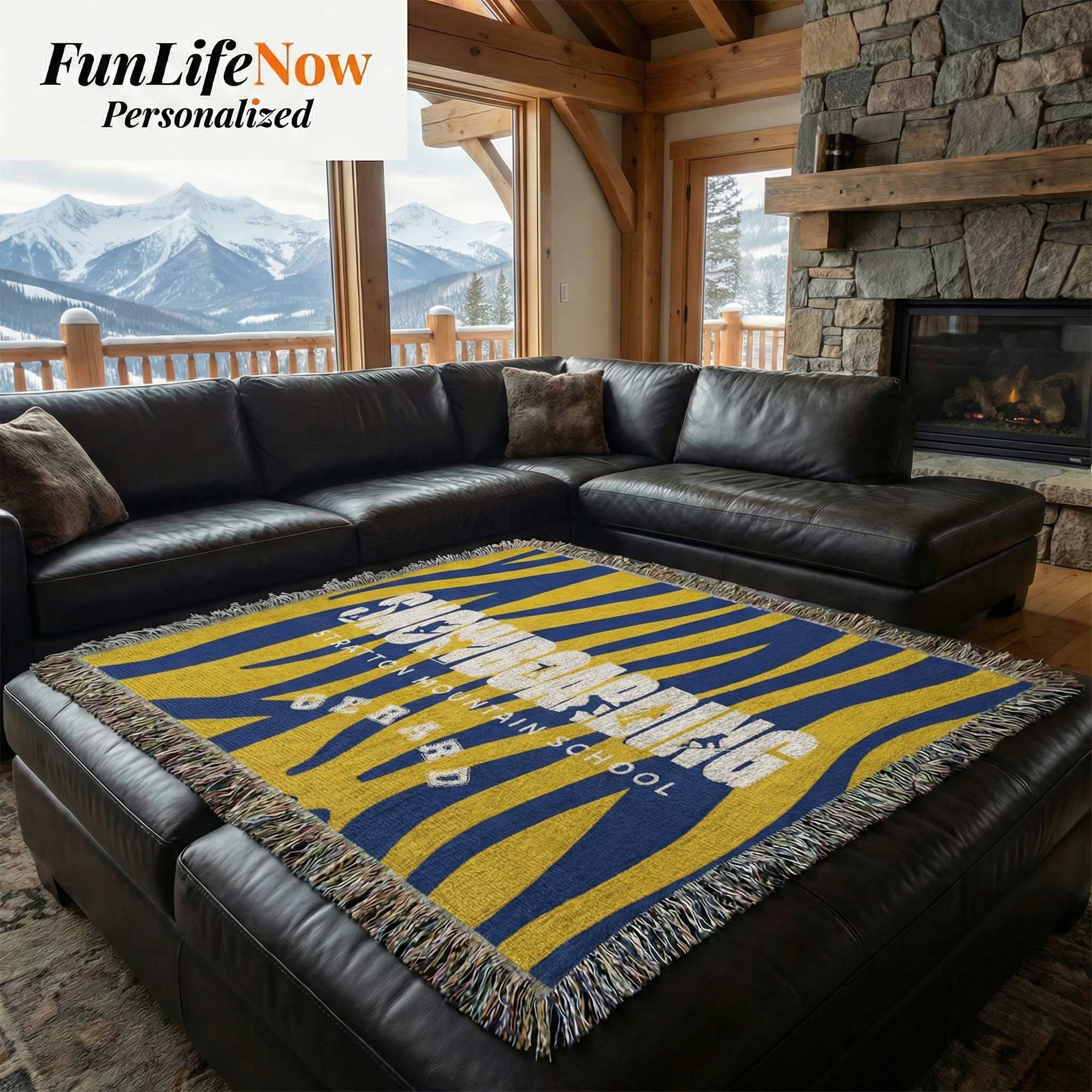 Black leather sectional sofa with a personalized snowboard blue and yellow patterned throw blanket in a room with a stone fireplace and mountain view. Custom blanket available at Funlifenow.com
