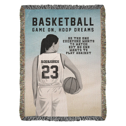 A personalized woven blanket with a silhouette of a girl in a basketball uniform holding a basketball, with the text 'BASKETBALL GAME ON, HOOP DREAMS' and a name and jersey number which are customizable.
